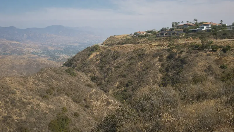 Los Angeles County, California: Steep chaparral canyon (chamise, sage); modern homes along a winding ridge road; dry brush, haze