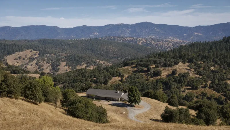 Placer County, California: Golden grass hills with blue oaks turning into pine forest; single-story ranch house with gravel drive and mowed strip