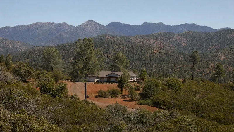 Shasta County, California: Red-soil hills with manzanita, scrub oak and gray pine; rural single-story home on a big lot; forested ridges behind